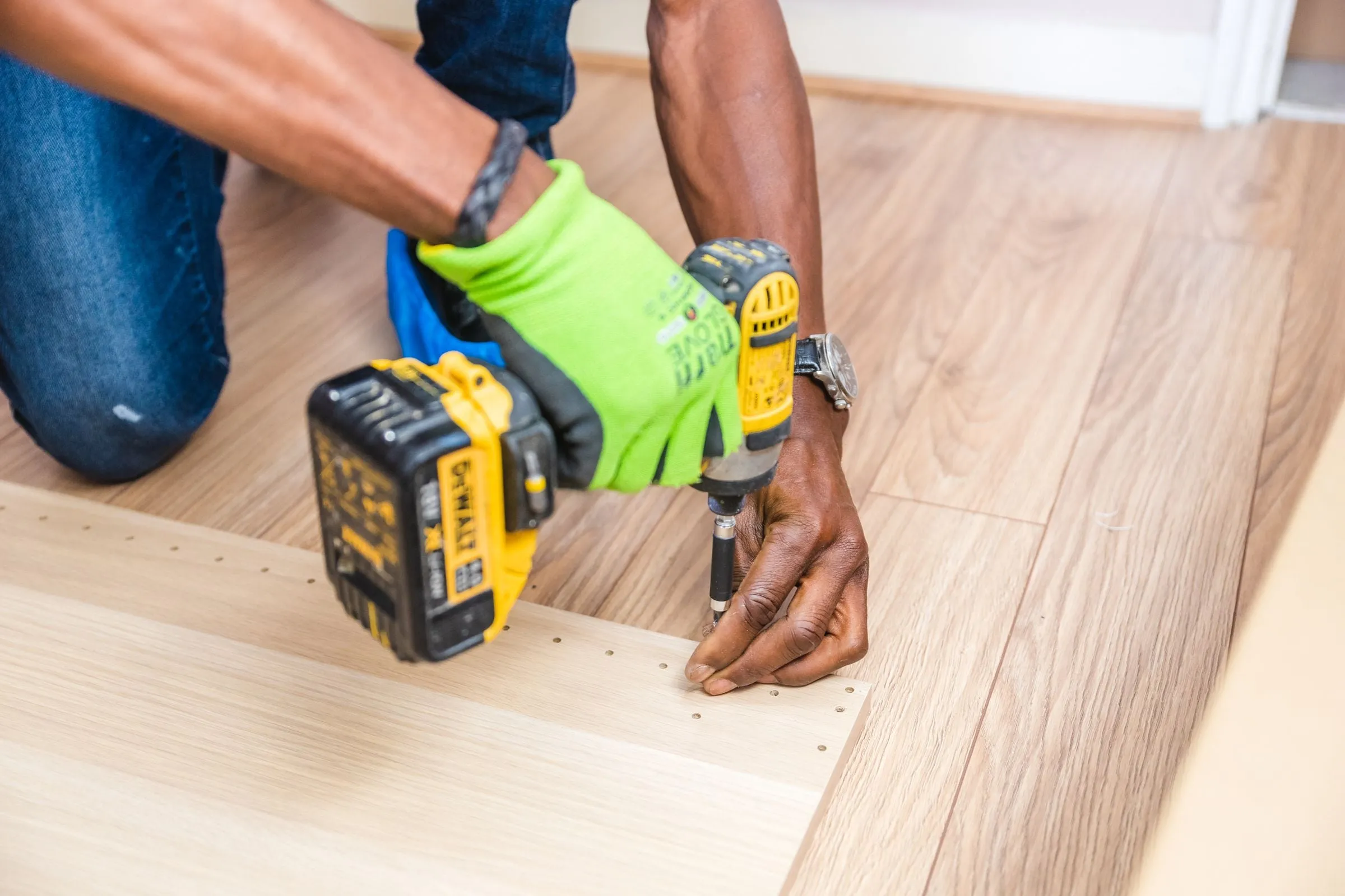 An installer in work gloves driving screws into a furniture panel with a cordless drill