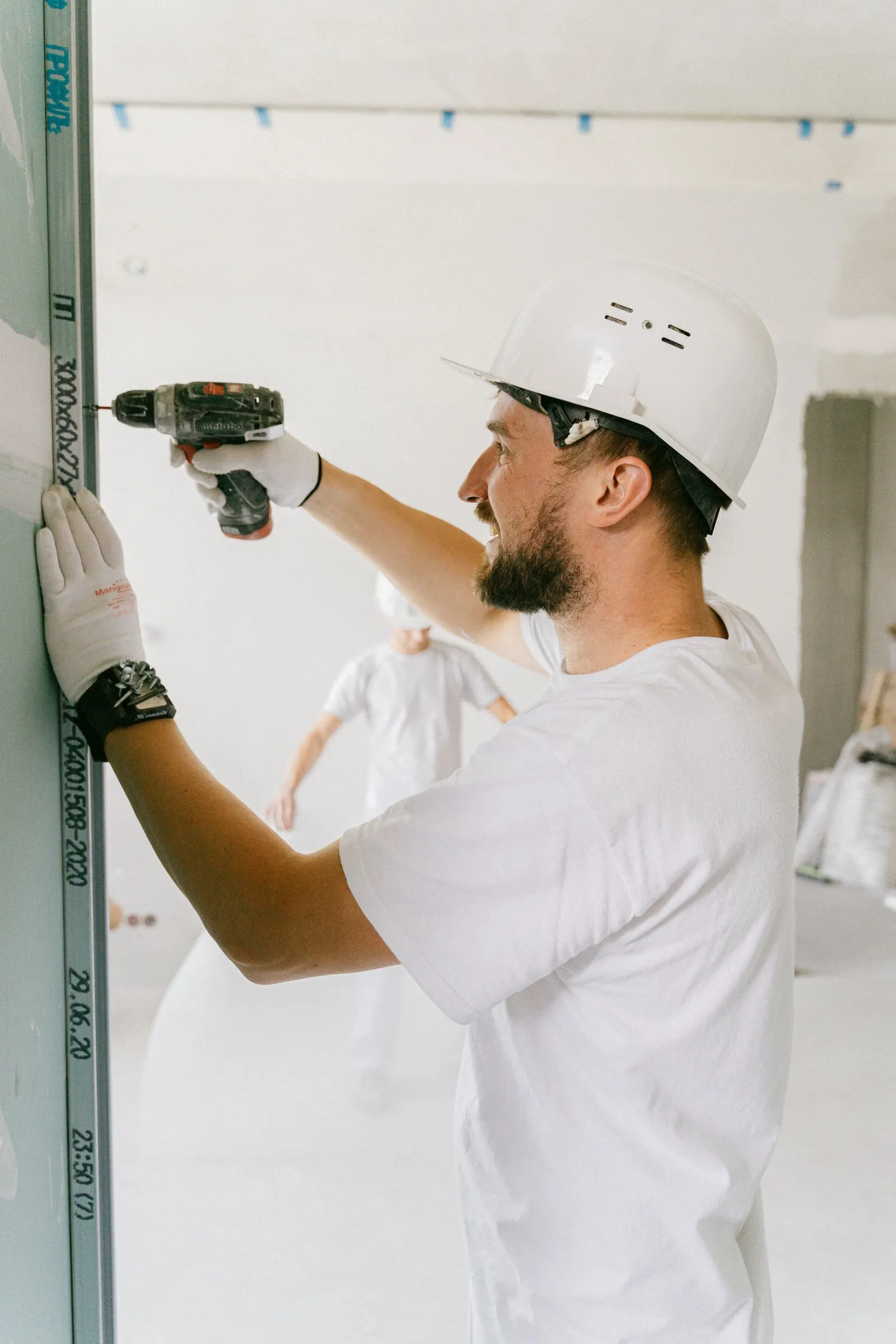 An installer in a hard hat and gloves using a drill and level on a wall